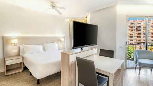 Valoisa makuuhuone, jossa parivuode, pöytä tuoleineen ja suuri televisio sängyn edessä. (Tekoälyn luoma teksti)