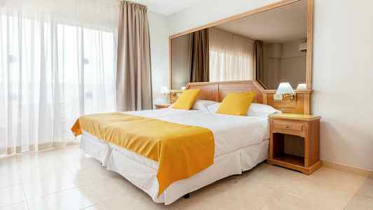 Hotellihuone, jossa on parivuode, valkoiset ja keltaiset vuodevaatteet. Sängyn takana on suuri peili ja ikkunassa vaaleat verhot. (Tekoälyn luoma teksti)