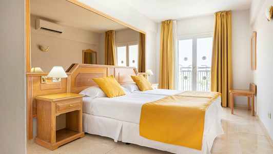 Hotellihuone, jossa on parivuode, valkoiset lakanat ja keltaiset tyynyt ja päiväpeite. Huoneessa on vaaleat puuhuonekalut ja ikkunat verhoineen. (Tekoälyn luoma teksti)