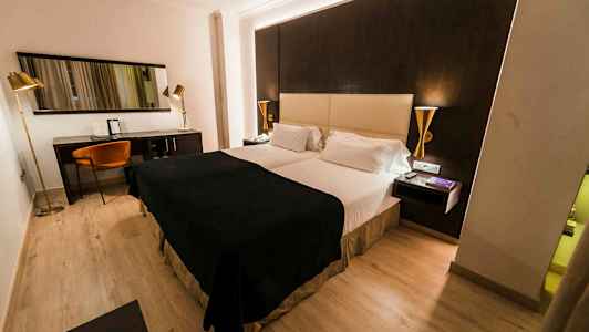 Hotellihuone, jossa on parisänky, tumma ja vaalea värisävy huonekaluissa sekä työpiste tuolin ja peilin kanssa. (Tekoälyn luoma teksti)