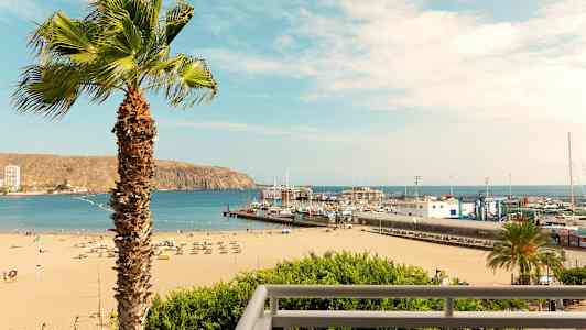 Ranta aurinkotuoleineen ja aurinkovarjoineen, satama veneineen ja palmu etualalla. (Tekoälyn luoma teksti)