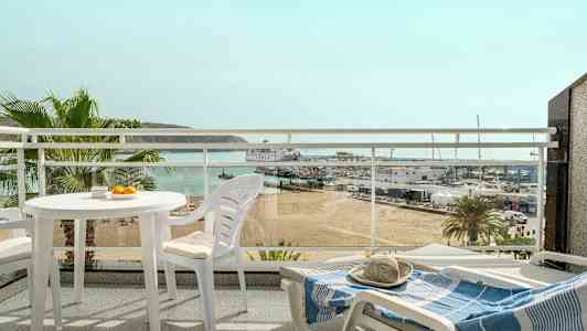 Näkymä parvekkeelta, jolla on valkoiset ulkokalusteet, näkymänä ranta, pienvenesatama ja meri taustalla. (Tekoälyn luoma teksti)