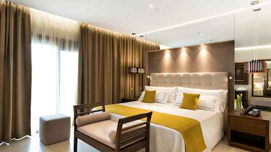 Hotellihuone, jossa on suuri sänky, vaaleat seinät ja verhot sekä istuinpenkki jalkopäässä. (Tekoälyn luoma teksti)