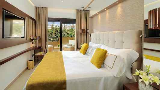 Moderni hotellihuone, jossa on leveä sänky, koristeltu keltaisilla yksityiskohdilla ja näkymä parvekkeelle. (Tekoälyn luoma teksti)