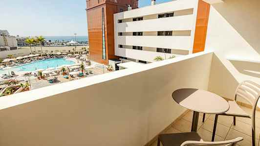 Näkymä parvekkeelta kohti allasaluetta, jossa on aurinkotuoleja, aurinkovarjoja ja palmuja, sekä valkoisia ja punaisia rakennuksia ja meri taustalla. Kaksi tuolia ja pöytä näkyvät parvekkeella. (Tekoälyn luoma teksti)