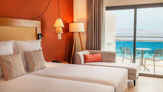 Hotellihuone, jossa on parivuode, koristeellisia tyynyjä, sohva ja lasiovet, jotka johtavat parvekkeelle merinäkymällä. (Tekoälyn luoma teksti)