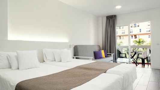 Hotellihuone, jossa on kaksi erillistä sänkyä valkoisin vuodevaattein, harmaa sohva värillisillä tyynyillä ja parveke näkymällä. (Tekoälyn luoma teksti)