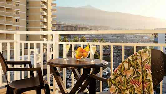 Parveke, jossa on pöytä, kaksi lasia, kulho hedelmiä ja ympärillä näkymät ympäröiville rakennuksille ja vuorille. (Tekoälyn luoma teksti)