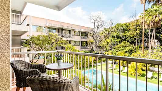 Näkymä parvekkeelta, jolla on kaksi rottinkituolia pienen pöydän ääressä, uima-allas ja viheralueita sekä asuinrakennus taustalla. (Tekoälyn luoma teksti)