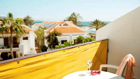 Näköala parvekkeelta, jossa on keltainen kaiteen suoja, pieni pöytä, jossa lasi ja punaiset aurinkolasit, valkoinen tuoli sekä lomamökit ja meri taustalla. (Tekoälyn luoma teksti)