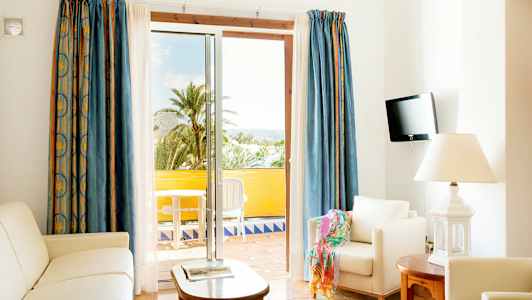 Valoisa olohuone, jossa on valkoiset huonekalut ja avoin parvekkeen ovi näkymällä palmuihin. (Tekoälyn luoma teksti)