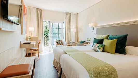 Vaalea hotellihuone, jossa on kaksi vuodetta, koristeelliset tyynyt ja työskentelytila parvekkeen vieressä. (Tekoälyn luoma teksti)