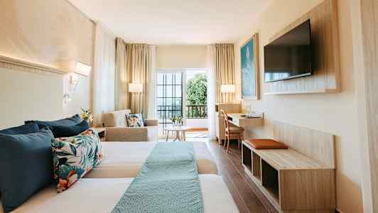 Hotellihuone, jossa kaksi sänkyä, sohva, pöytä ja televisio sekä parveke lasiovilla. (Tekoälyn luoma teksti)