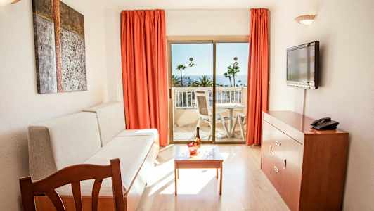 Huone, jossa on sohva, pöytä, televisio ja parveke, josta avautuu näkymä palmuihin ja merelle. (Tekoälyn luoma teksti)