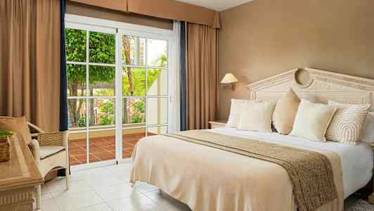 Hotellihuone, jossa on suuri sänky, beiget vuodevaatteet, valkoiset tyynyt ja lasiovi, joka avautuu terassille, jolla on vihreää kasvillisuutta. (Tekoälyn luoma teksti)