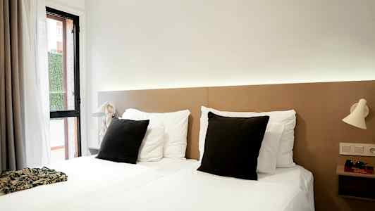 Hotellihuone, jossa on kaksi erillistä sänkyä, valkoiset vuodevaatteet ja mustat tyynyt sekä vaaleat yöpöytälamput seinällä. (Tekoälyn luoma teksti)