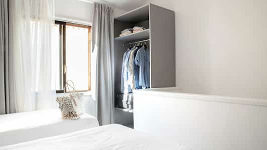 Huone, jossa on kaksi yhden hengen vuodetta, verhot ikkunassa ja avoin vaatekaappi vaatteineen. (Tekoälyn luoma teksti)