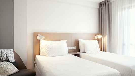 Hotellihuone, jossa on kaksi erillistä vuodetta, verhoilla varustettu ikkuna ja tuoli sivussa. (Tekoälyn luoma teksti)