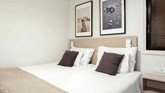 Makuuhuone, jossa on kaksi yhden hengen sänkyä, valkoiset lakanat, tummat tyynyt ja kaksi kehystettyä kuvaa seinällä. (Tekoälyn luoma teksti)