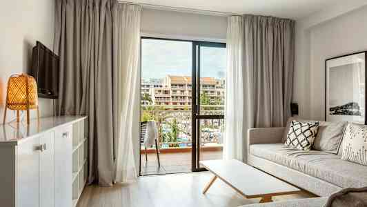 Valoisa olohuone, jossa on sohva, pöytä ja näkymä parvekkeelle sekä taustalla rakennuksia. (Tekoälyn luoma teksti)