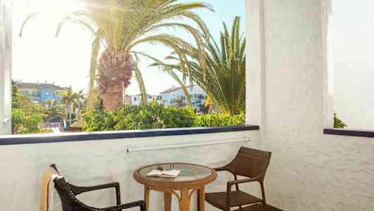 Parveke, jossa on kaksi tuolia ja pöytä kirjan kanssa. Parvekkeen takana näkyy palmu ja rakennuksia. (Tekoälyn luoma teksti)