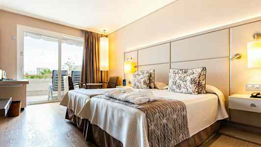 Hotellihuone, jossa kaksi sänkyä ja neutraalejä sävyjä. Näkymä parvekkeelle lasiovien kautta. (Tekoälyn luoma teksti)