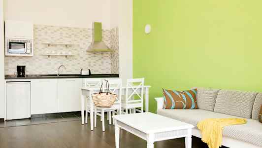Olohuone, jossa on vihreät seinät, sohva tyynyillä ja pöytä sekä viereinen keittiö, jossa valkoiset kaapit ja kodinkoneet. (Tekoälyn luoma teksti)
