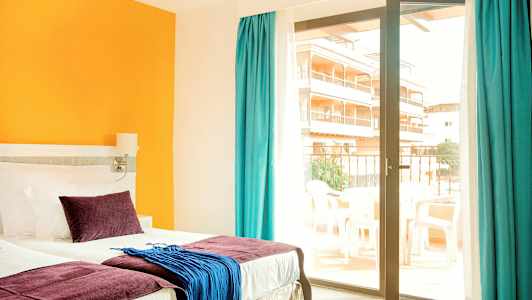 Hotellihuone, jossa on sänky, värikkäitä tyynyjä ja tekstiilejä sekä lasiovi, joka johtaa parvekkeelle. (Tekoälyn luoma teksti)