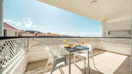 Parveke, jossa pöytä ja tuolit, näkymä ympäröiviin rakennuksiin ja vuoristoon. (Tekoälyn luoma teksti)