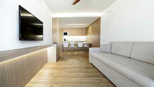 Moderni olohuone, jossa vaaleat sisustukset, harmaa sohva, seinään asennettu televisio ja keittiö taustalla. (Tekoälyn luoma teksti)
