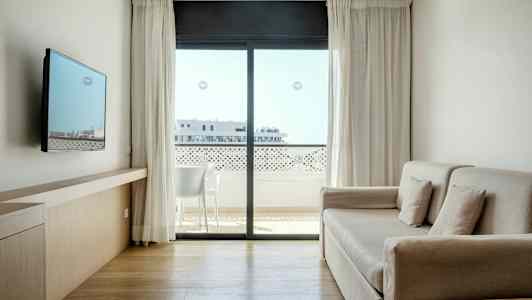 Valoisa ja moderni hotellihuone, jossa on sohva, seinälle kiinnitetty TV ja suuri ikkuna, josta avautuu näkymä parvekkeelle. (Tekoälyn luoma teksti)