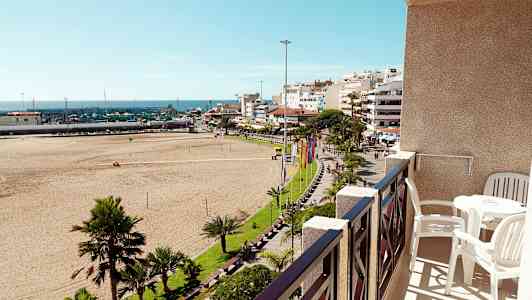 Näkymä parvekkeelta, jossa on pöytä ja tuolit, kohti hiekkarantaa, vehreää puistoa ja kaupunkimaisemaa. (Tekoälyn luoma teksti)