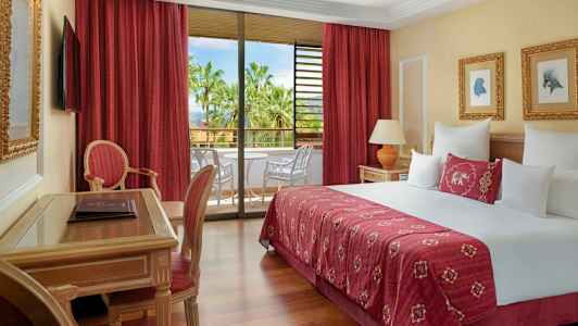 Hotellihuone, jossa on suuri sänky, istumakalusteet ja parveke näkymällä palmuihin. (Tekoälyn luoma teksti)