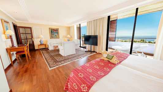 Tilava hotellihuone, jossa parivuode, istuinryhmä, kirjoituspöytä ja parveke maisemilla. (Tekoälyn luoma teksti)