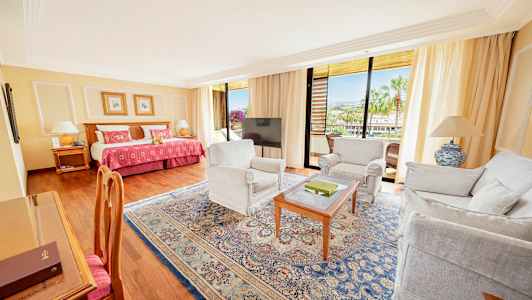 Hotellihuone, jossa on sänky, istuinryhmä ja parveke näkymineen. (Tekoälyn luoma teksti)