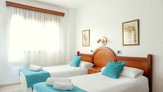 Kaksi yksittäistä sänkyä, joissa turkoosit koriste-tyynyt ja valkoinen päiväpeite, aurinkoisessa huoneessa, jossa iso ikkuna. (Tekoälyn luoma teksti)