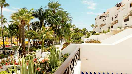 Näkymä puutarhaan, jossa on useita huojuvia palmupuita ja valkoisia rakennuksia, joissa on terasseja. (Tekoälyn luoma teksti)