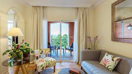 Olohuone, jossa on koristeellinen tuoli, sohva ja kukkia, näkymä parvekkeelle. (Tekoälyn luoma teksti)