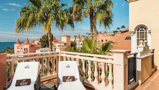 Kaksi aurinkotuolia terassilla, näkymä palmuihin ja pastellinvärisiin rakennuksiin. (Tekoälyn luoma teksti)