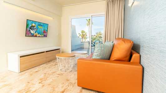 Moderni olohuone, jossa oranssi sohva, tyynyjä, sohvapöytä ja TV, näkymä parvekkeelle ja palmumetsään. (Tekoälyn luoma teksti)