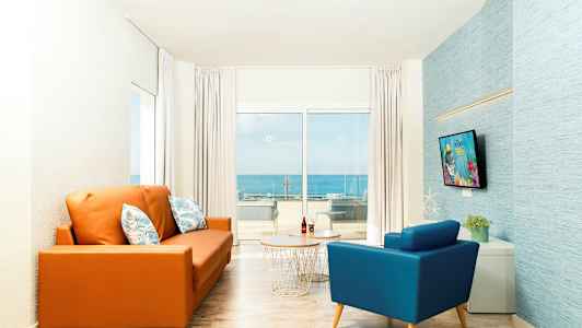 Olohuone, jossa on oranssi sohva ja sininen nojatuoli pöydän edessä. Iso ikkuna taustalla näyttää näkymän merelle. (Tekoälyn luoma teksti)