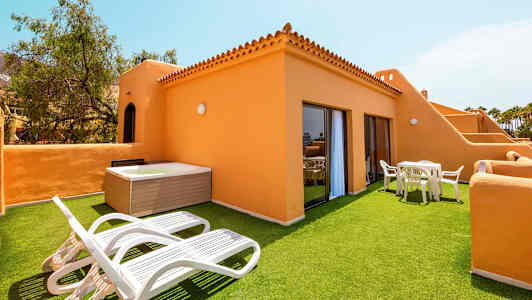 Terassi tekonurmella, aurinkotuoleilla, pöydällä ja tuoleilla sekä porealtaalla, oranssien rakennusten ympäröimänä. (Tekoälyn luoma teksti)