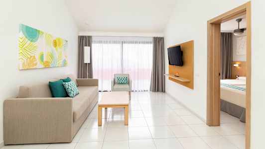 Hotellihuone, jossa näkyy osittain vuode, sohva, nojatuoli ja pöytä. Taustalla on ikkuna ja verhot. (Tekoälyn luoma teksti)