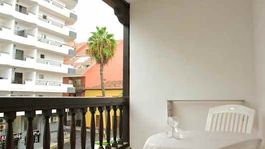 Parveke, jossa pöytä, kaksi lasia ja valkoinen muovituoli. Näkymä näyttää rakennuksia, palmupuun ja kadun. (Tekoälyn luoma teksti)