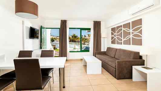 Valoisa ja modernisti sisustettu hotellihuone, jossa ruskea sohva, ruokapöytä ja tuolit. Ikkunat ja ovi johtavat ulkotilaan. (Tekoälyn luoma teksti)