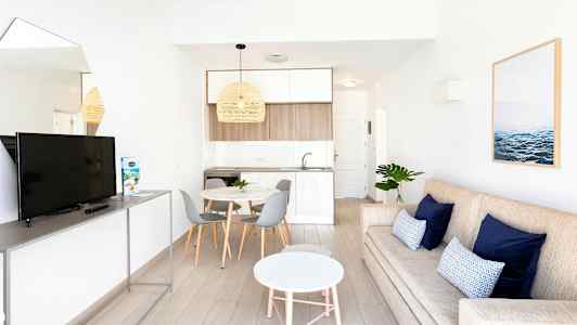 Moderni olohuone, jossa beige sohva, siniset tyynyt, valkoinen pyöreä sohvapöytä ja televisio jalustalla. Vieressä on ruokailutila, jossa pyöreä pöytä ja harmaat tuolit sekä vaaleansävyinen keittiö. (Tekoälyn luoma teksti)