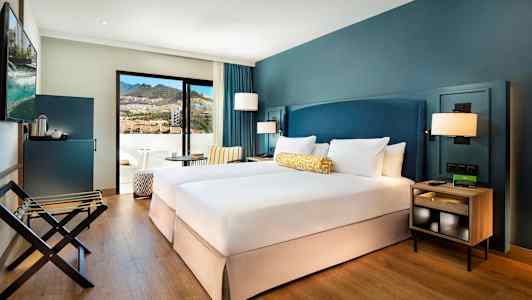 Hotellihuone, jossa on kaksi erillistä sänkyä, siniset seinät ja parveke vuorinäköalalla. (Tekoälyn luoma teksti)