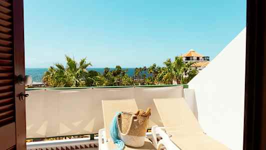 Näkymä aurinkoiselle parvekkeelle, jolla on aurinkotuoleja, korilaukku, palmuja ja meri taustalla. (Tekoälyn luoma teksti)