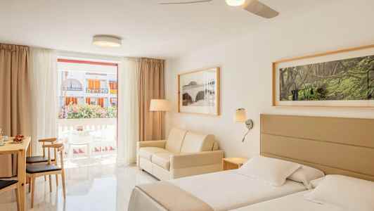 Valoisa hotellihuone, jossa on kaksi sänkyä, sohva ja ruokapöytä tuoleineen sekä suuri ikkuna näkymällä parvekkeelle ja ulkopuolella oleviin rakennuksiin. (Tekoälyn luoma teksti)