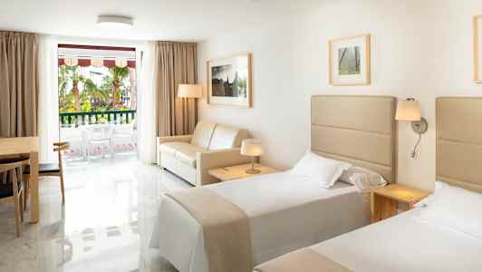 Hotellihuone, jossa on kaksi erillistä vuodetta, sohva, pöytä tuoleineen ja ikkuna parvekkeelle. (Tekoälyn luoma teksti)